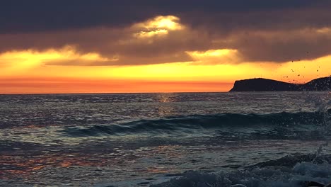 Golden-hour-on-seaside-with-orange-sky-and-dark-clouds-over-sea-and-silhouette-mountains,-dusk-light-reflecting-on-sea-water