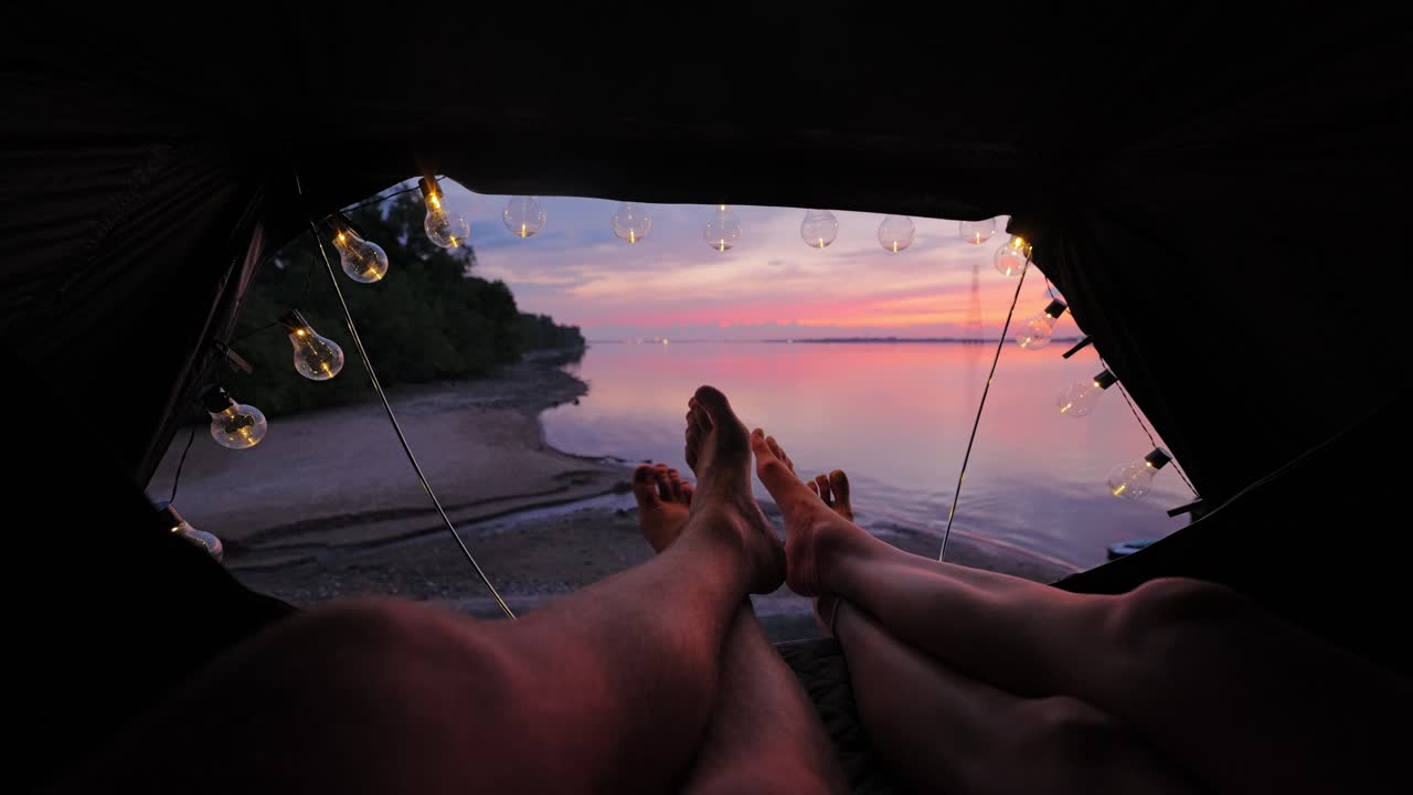 Two people relax, rooftop tent at sunset, warm bulb lights, romantic river view