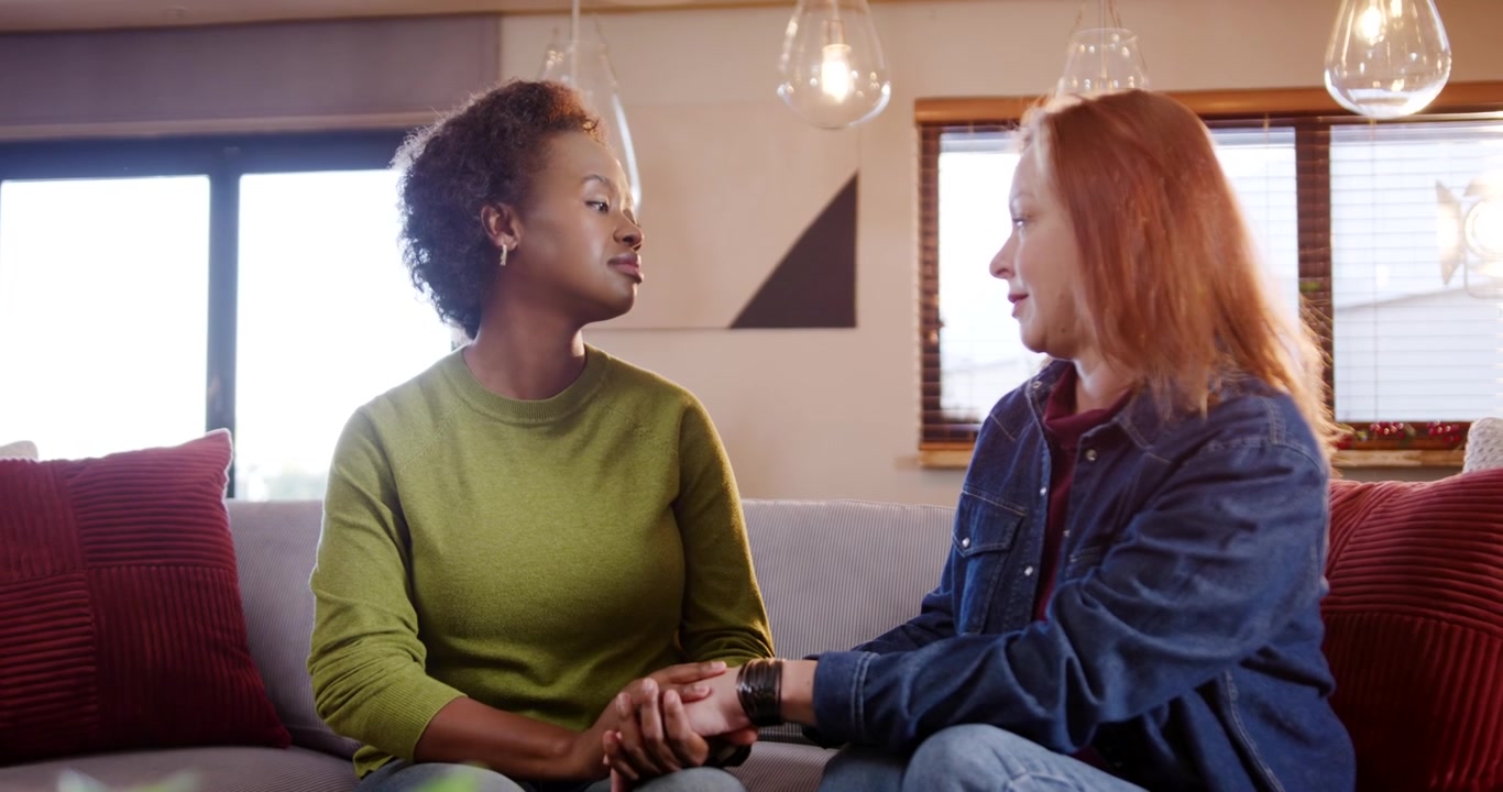 Two female friends sitting on gray couch, red-haired friend reaching for mug and comforting other