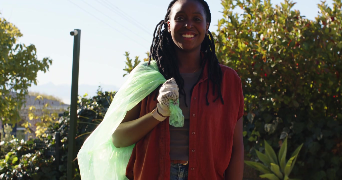 African woman carrying green bag wearing gloves holding tied plastic during cleanup on lawn