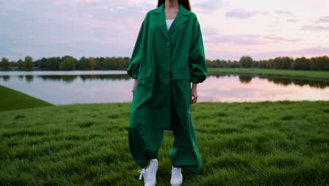 woman in green coat and white sneakers outdoors by a lake at sunset