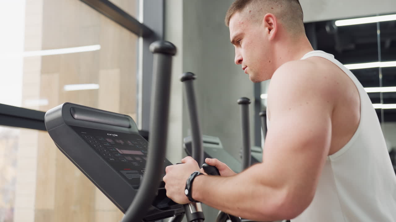 Young man fixed eyes on gym equipment console during intense focus workout in modern studio with floor to ceiling glass windows reflecting action and dynamic athletic posture of determination