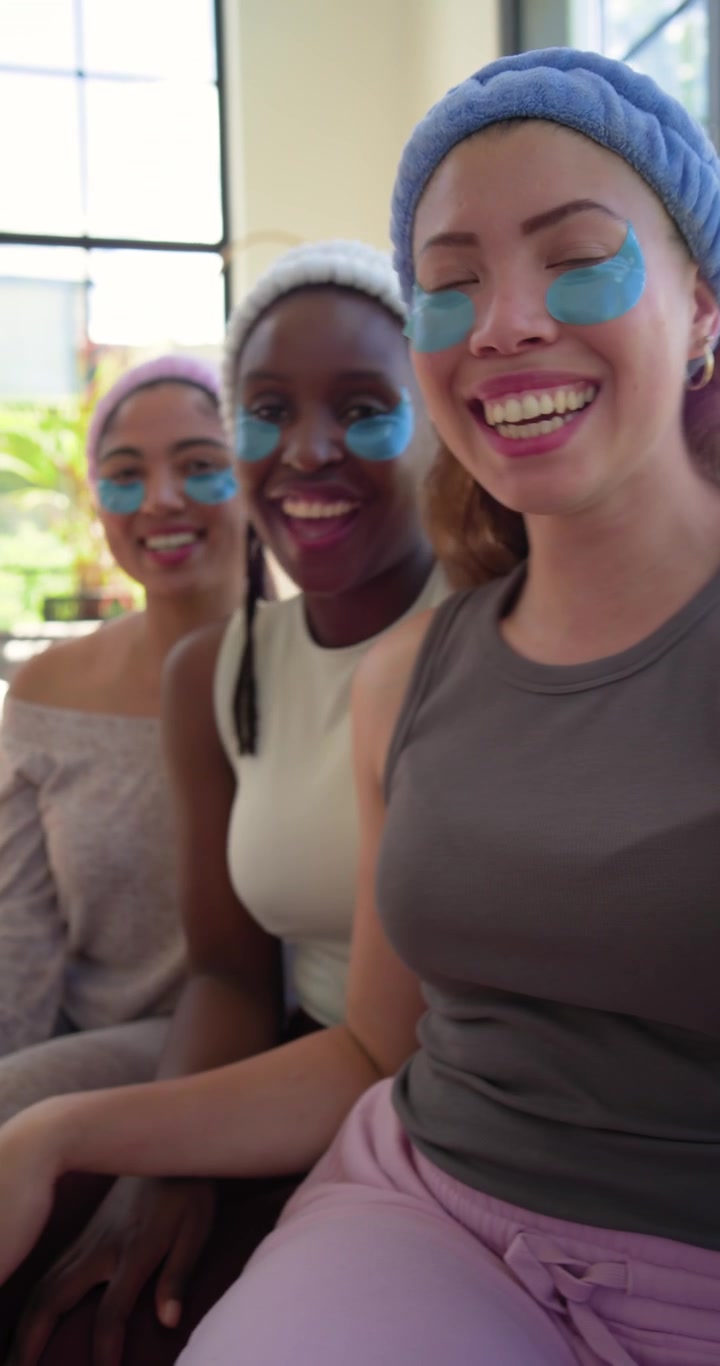 Video verticale: Tre donne che ridono sul divano con maschere blu, fasce per capelli, che filmano un selfie con il telefono