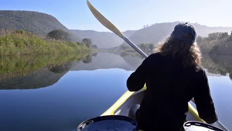 Girl-canoeing-over-mirror-smooth-river-with-hazy-hills-in-the-background-in-Wilderness-National-Park,-South-Africa