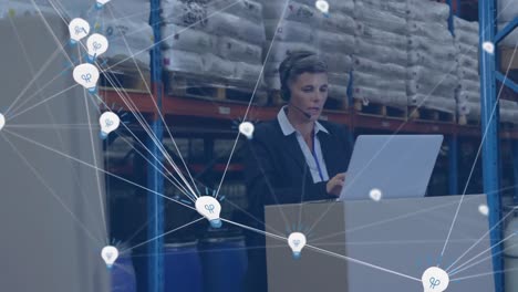 animation of network of bulb icons over caucasian female supervisor using laptop at warehouse