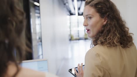 Diverse-female-colleagues-in-discussion-using-laptop-and-tablet-in-office-corridor,-slow-motion