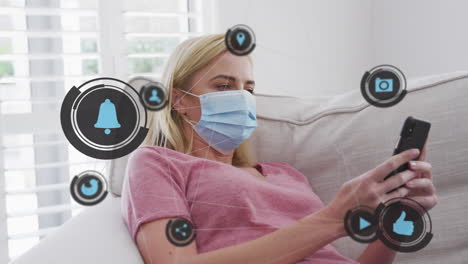 animation of digital icons over caucasian woman in face mask using smartphone at home