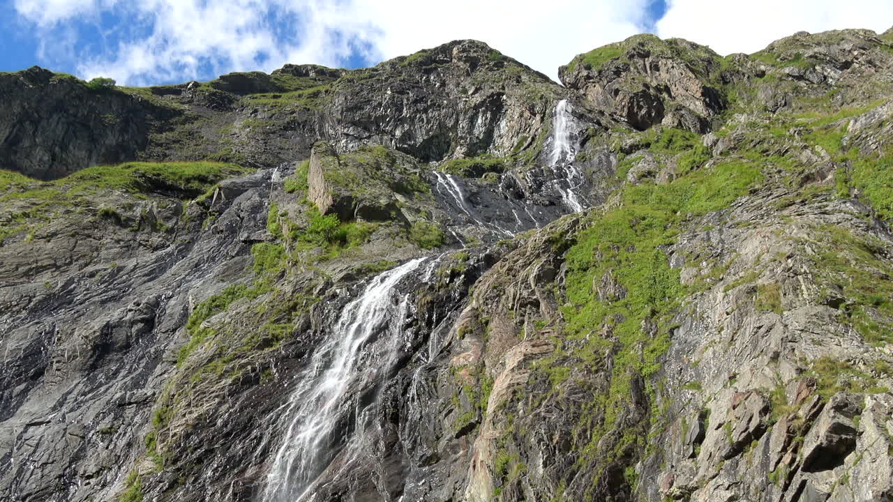 Suggestiva cascata di montagna che scende da ripide scogliere rocciose con vegetazione alpina verde sotto un cielo nuvoloso a Dombay, Caucaso, Russia, ideale per temi di viaggio e natura.