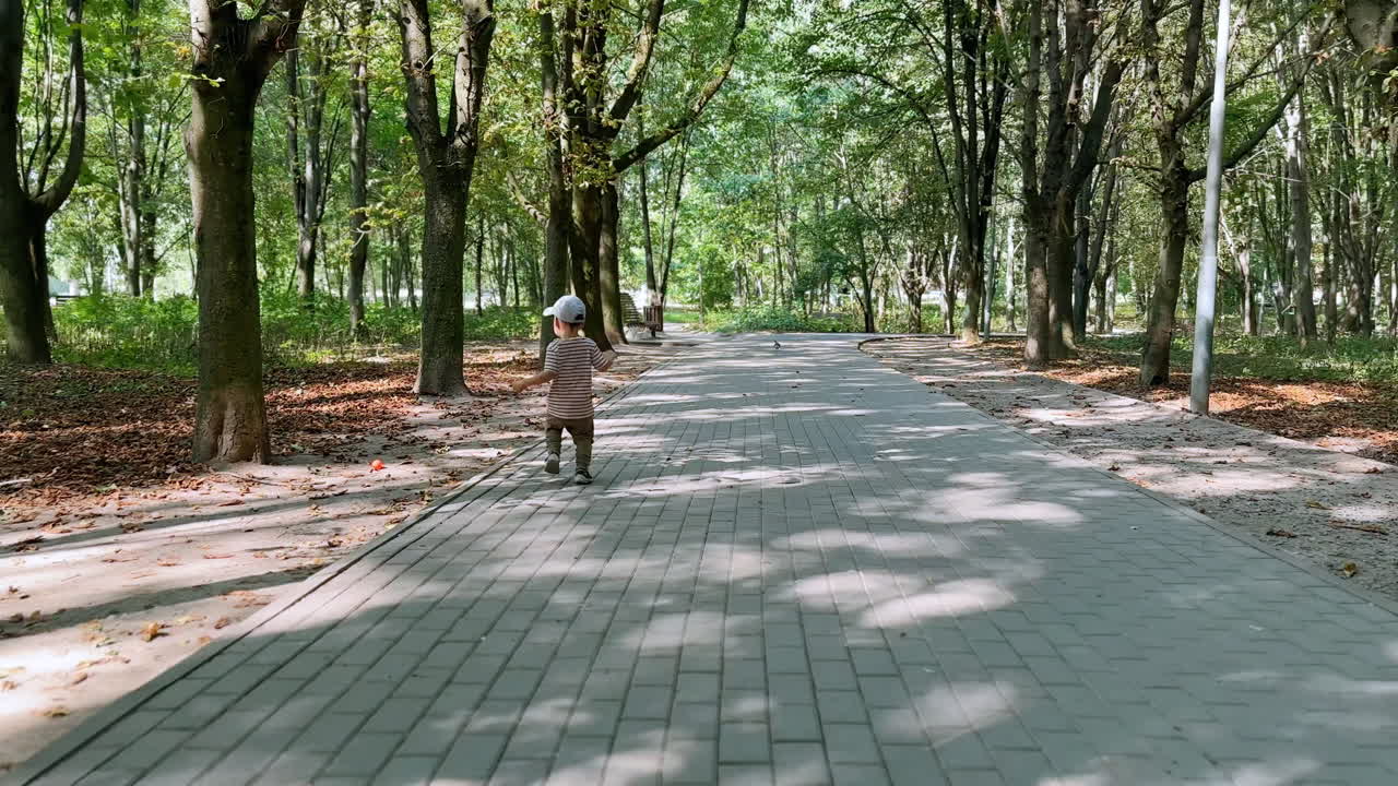 Rear view of a toddler boy running by the tiled path in the park. Happy kid picks up a ball and throws it again.