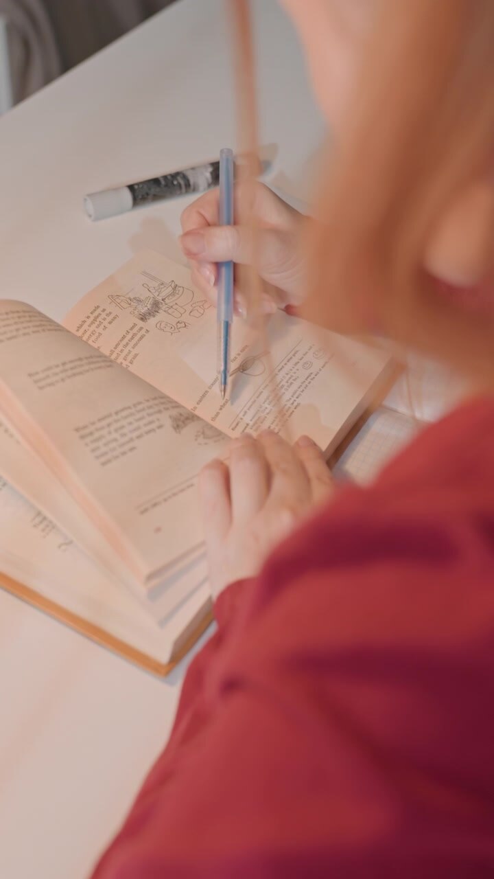 Young woman with light brown hair wearing red top reads. Woman uses her right hand to point or underline text. Scene is well lit indoors with soft lighting focusing closely