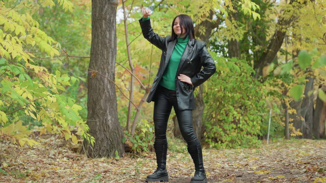 Black woman in leather jacket dancing through autumn forest with green sweater and tall boots, striking confident poses and playful interaction with tree branch, candid motion across textured fallen