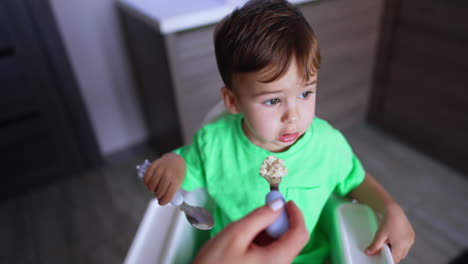 mom is holding a full fork in front of her little son. toddler refuses from food but then takes it. top view.
