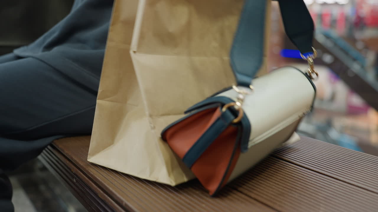 Close up of shopper seating down beside brown bench inside brightly lit shopping mall, holding paper bag and stylish handbag with orange and white design, blurred background