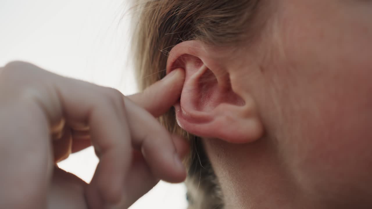 Primo piano di ragazza adolescente bianca che si tocca l'orecchio, tramonto all'aperto, dito che preme il lobo, controlla cerume e pressione dopo la corsa, profilo spontaneo con acne e pelle lentigginosa, delicata routine di cura personale, luce dorata e soffusa.