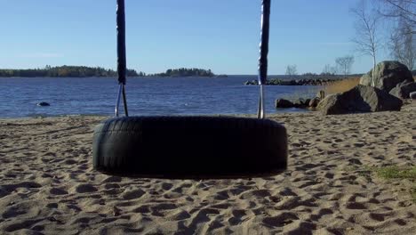 Neumático-De-Camión-Reciclado-Transformado-En-Un-Columpio-De-Juegos-En-La-Playa,-Balanceándose-Lentamente-De-Un-Lado-A-Otro-Con-El-Mar-Y-La-Playa-En-El-Fondo,-Columpio-De-Neumáticos-De-Goma-Sin-Uso,-Era-Digital-Y-Parques-Infantiles-Vacíos