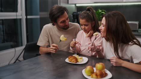 A-happy-brunette-man-with-stubble-in-a-beige-T-shirt-together-with-his-brunette-wife-in-a-white-T-shirt-and-a-little-daughter-in-a-pink-suit-eat-cheesecakes-during-their-breakfast-in-a-modern-apartment-at-a-black-dining-table-in-the-kitchen-in-the-morning