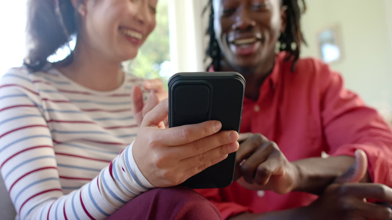 Happy diverse couple sitting on couch using smartphone in sunny living room, slow motion