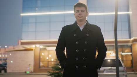 mall owner in black coat standing in front of mall with hand in pocket, sporting funny look, background features street lights on and parked cars, casual and lighthearted expression