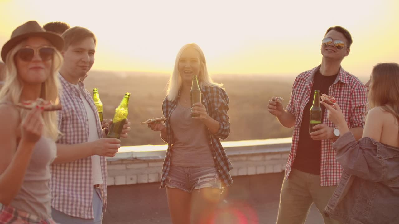 The company of six young persons having a party on the roof. They eat hot pizza and drink beer from the green bottles they smiles on a summer evening. They dance and communicate with each other.