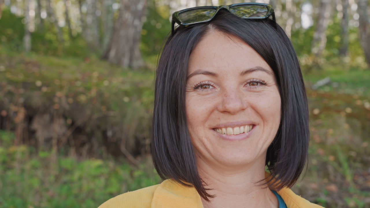 Primer plano de una mujer blanca acampando hablando a la cámara con expresiones juguetonas y ojos brillantes, gafas de sol apoyadas en la cabeza, chaqueta amarilla informal, fondo de abedules, narradora espontánea compartiendo consejos de búsqueda de alimentos.