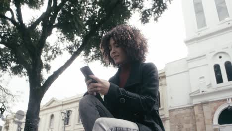 a curly woman in a suit sits in the park while replying to messages on her cellphone to finish work from her office