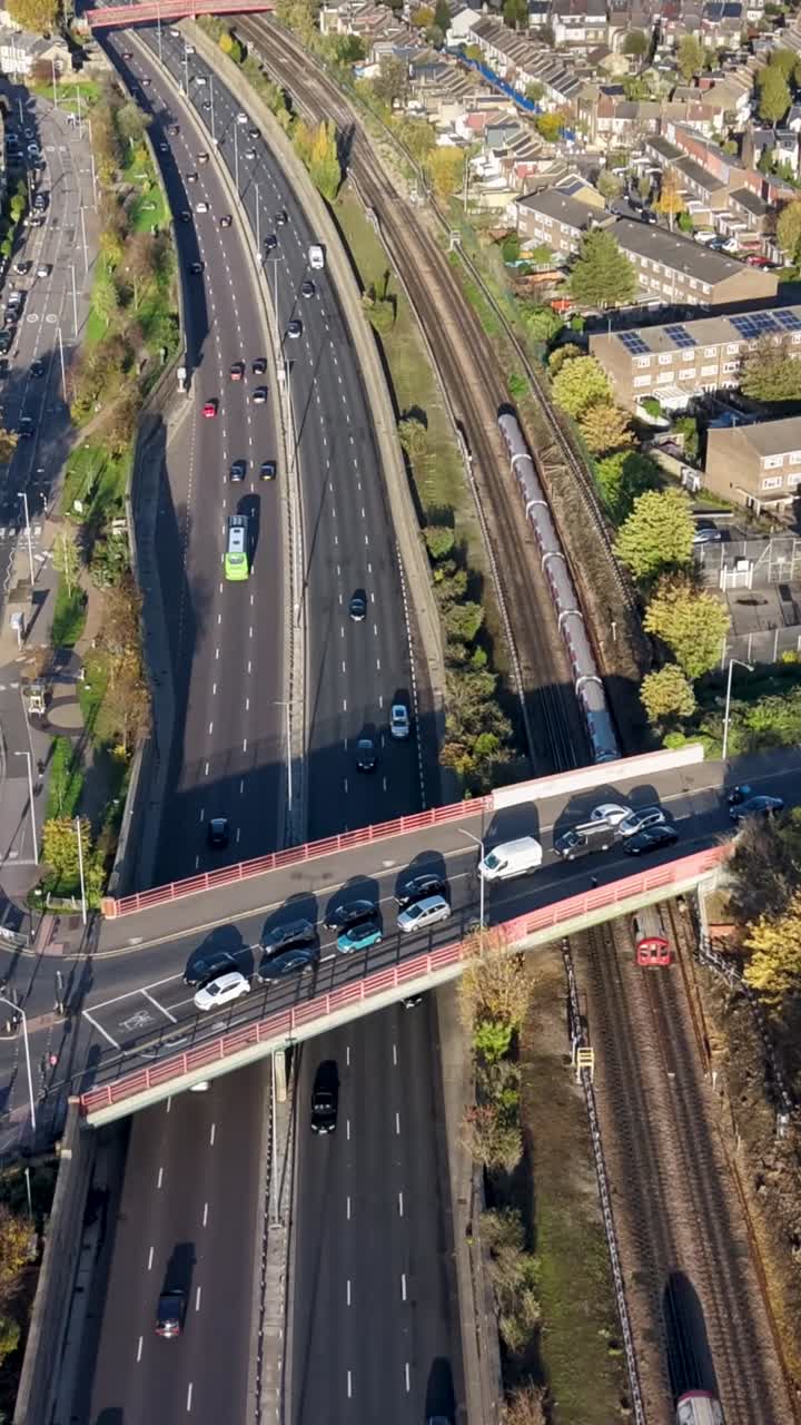 Vertical drone hyperlapse over A12 in Leytonstone, London, along road and Central Line train track, with cemetary to right. Cars and trains move below, with large cloud shadows drifting across