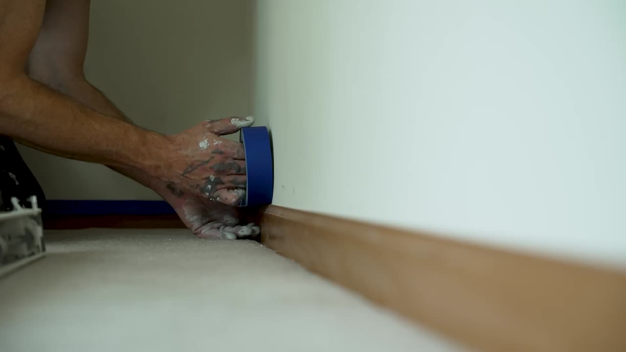 Een schilder brengt zorgvuldig blauwe afplaktape aan langs de plint om strakke randen voor te bereiden voor een nieuwe verflaag binnen, waarmee hij gedetailleerd vakmanschap en het precieze werk achter woningrenovatie laat zien