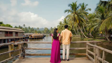 indian couple holding hands while admiring traditional houseboats along a tropical river on a sunny summer day, surrounded by lush palm trees and serene landscapes