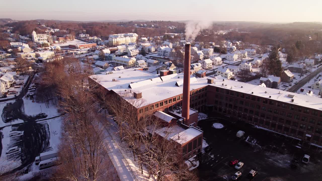 A tall brick smokestack stands over a snow covered town in early winter morning light