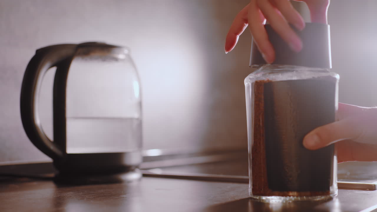 Close up of hands opening coffee container on kitchen counter with kettle in background, preparing to scoop coffee granules into cup during morning routine under warm natural light