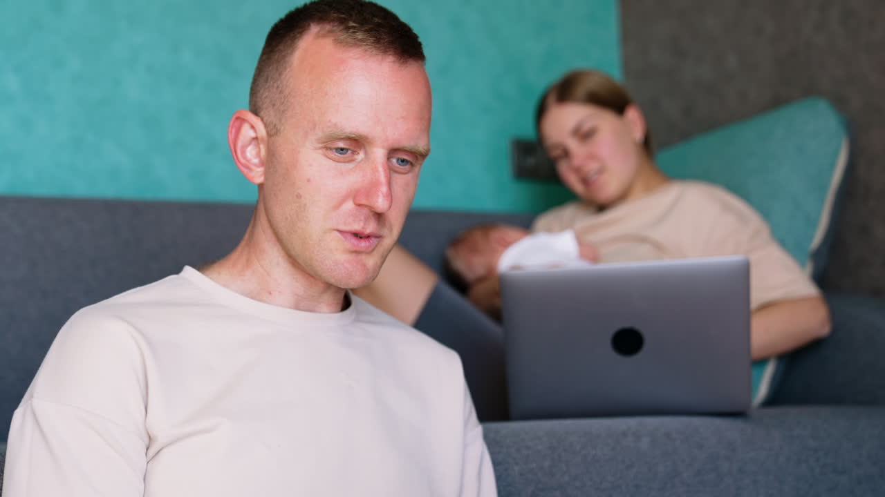 Caucasian man sitting on the floor focused on her laptop. Woman on sofa holding her baby looking at her laptop and talking to husband.