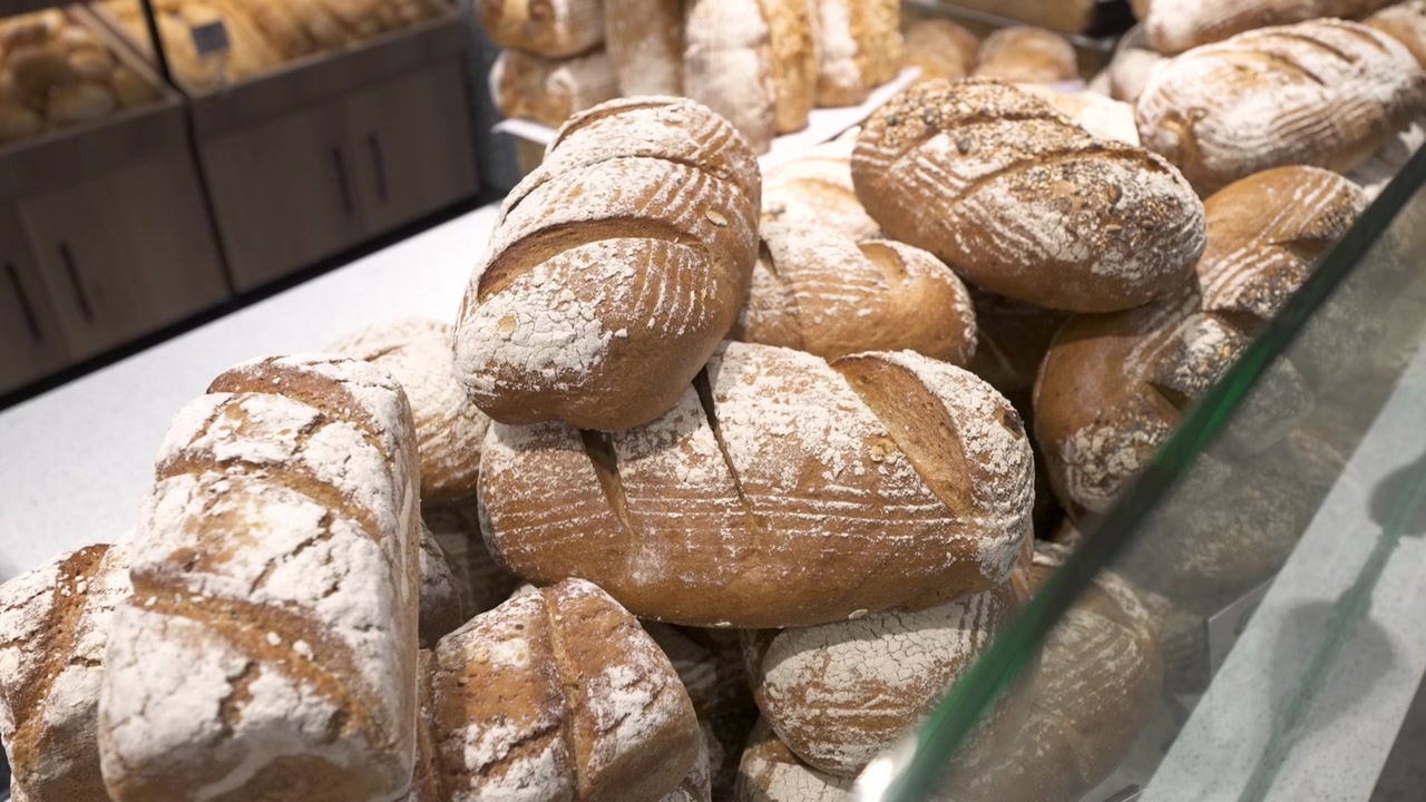 Crusty flour-dusted artisan loaves are stacked together behind glass in a retail food store bakery section.