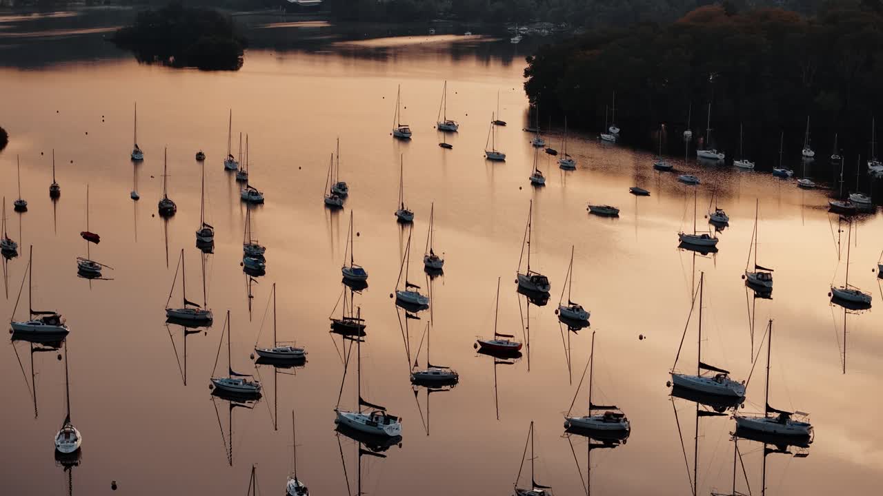 Aerial panning up along sailboats anchored on Windermere Lake, Lake District, England with stunning orange glow on water