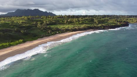 Aerial-view-over-clear-ocean-water-foaming-at-the-shore,-green-mountains