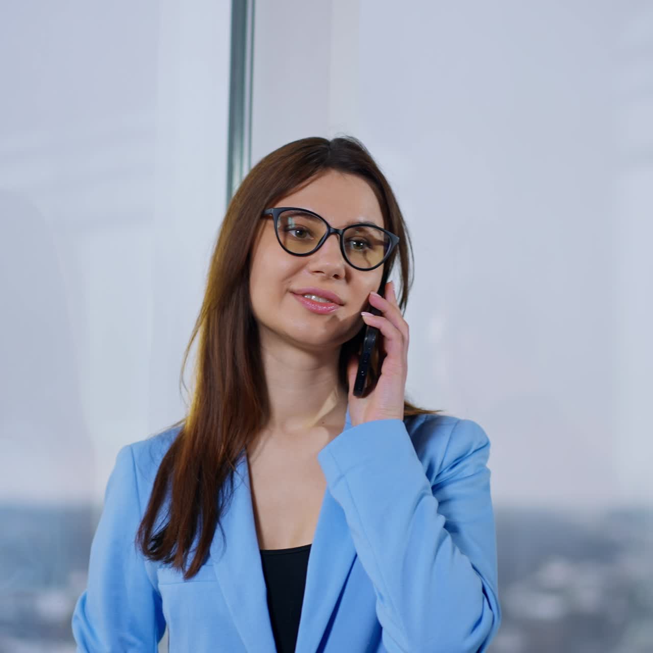 Brunette business lady is calling somebody on the phone. Man at backdrop using laptop standing at the window. Blurred background