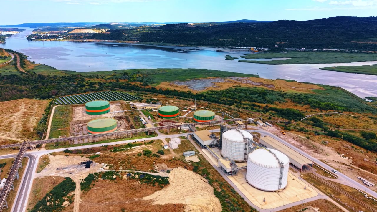 Serbatoi di stoccaggio accanto a zone umide fluviali e colline boscose. Cisterne bianche e verdi collegate da condotte vicino ad acque tranquille, canneti e binari ferroviari.