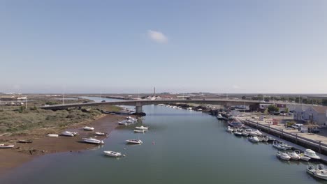 Aerial-pullback-from-bridge-over-Gilao-river,-Boats-moored-along-Riverbank,-Tavira