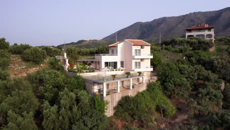 Aerial-Pull-Away-from-Luxury-Greek-Villa-with-Pool,-Sun-Loungers---Miniature-Palm-Trees-Revealing-Mountains-in-Background