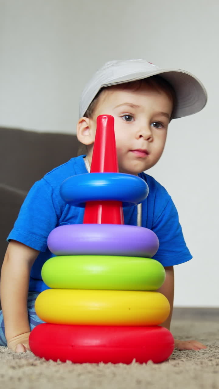 Smiling Caucasian baby boy comes up to a toy pyramid. Kid in a cap sits down on the floor to play with his toy. Low angle view. Vertical video