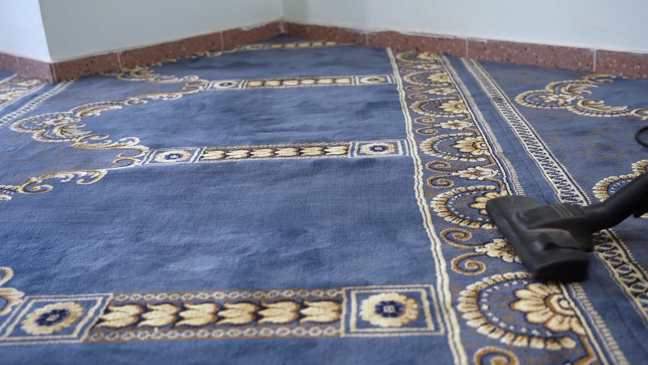 Primo piano di un ugello di aspirapolvere che pulisce un tappeto da preghiera blu a motivi in una moschea. Questa inquadratura evidenzia la manutenzione delle strutture religiose e la preparazione dello stile di vita islamico durante il giorno