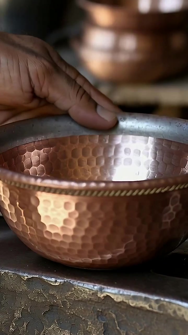Vertical video: Tilting adult male hand inspecting hammered copper bowl at bench, showing rope band