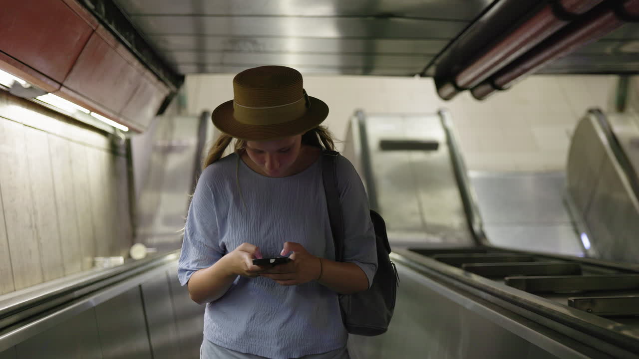 Jovem mulher usando smartphone em escada rolante, trajeto diário urbano e conceito de estilo de vida moderno.