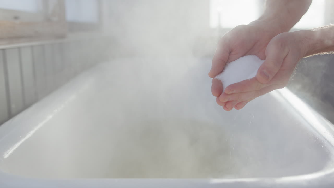 Cupped hands drop white epsom salts into steamy bath, telephoto