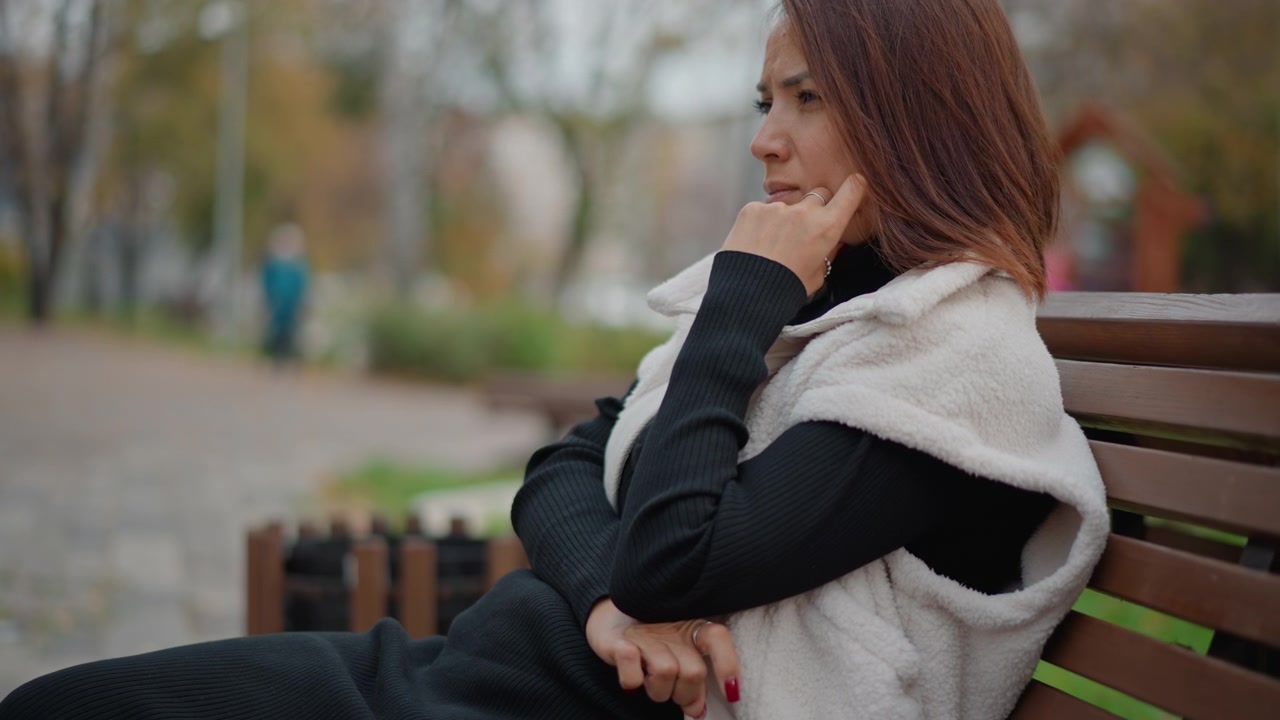 Donna adulta siede su una panchina di legno nel parco in una giornata autunnale dai colori tenui, chinata leggermente in avanti con una mano vicino al viso e le mani giunte in grembo, suggerendo attesa silenziosa, riflessione e calma