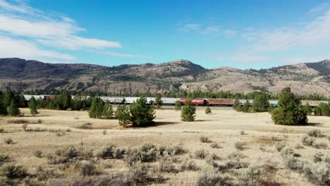 dolly forward drone shot flying towards a railroad station in a desert environment on a sunny day with mountains and trees in the background and a bush and grassland in the foreground