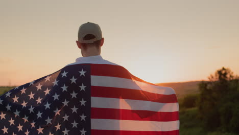 joven granjero con la bandera de estados unidos en los hombros mira el amanecer sobre el campo de trigo