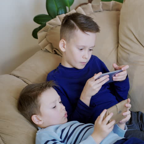 two cute boys relaxing on the couch and playing on his smaptphones at home. children spending time together holding phones in hands and playing competitive video games.