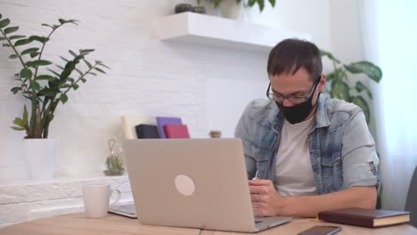 joven caucásico se queda en casa trabajando desde casa usando una máscara protectora usando computadora portátil e internet. oficina en casa acogedora, lugar de trabajo pandemia de coronavirus, cuarentena de covid-19. trabajo remoto, freelance