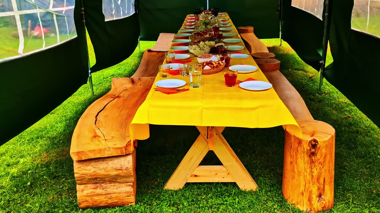 Long wooden picnic table set with plates and food under tent on bright green grass