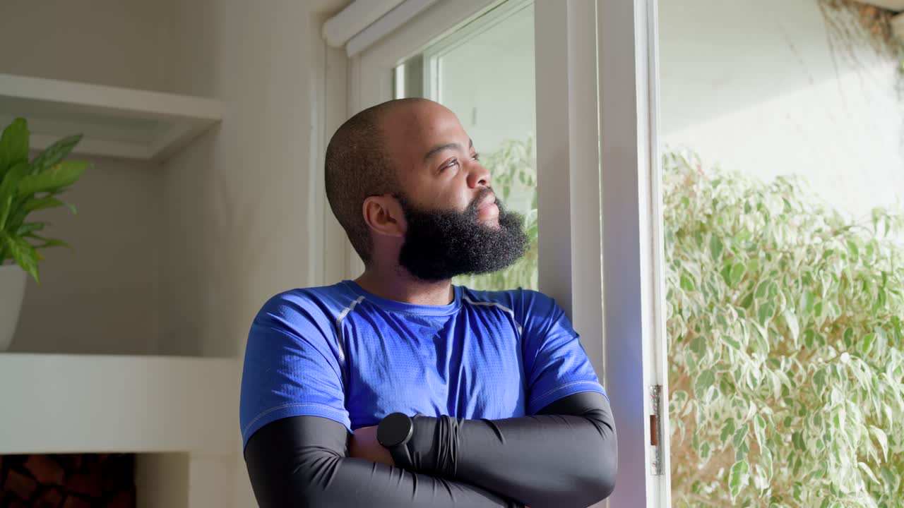 Uomo afroamericano in piedi davanti a una porta scorrevole a casa, con una camicia blu e uno smartwatch, sorridendo al fogliame.